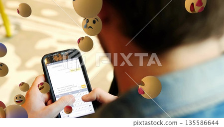 Typing blue-shirted man messaging on smartphone in cafe lounge, with floating yellow emoji spheres 135586644