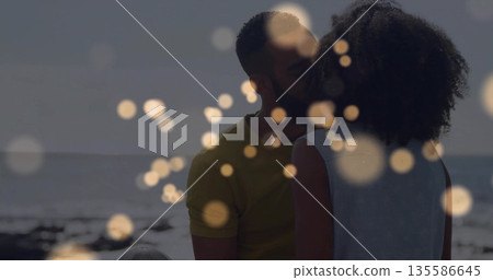 Kissing couple wearing summer attire on sandy shore, with ocean waves horizon line and bokeh lights 135586645
