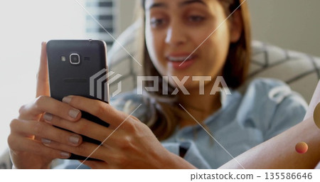 Smiling woman wearing blue top reclining on sofa near window blinds, holding black smartphone Smiling woman wearing blue top reclining on sofa near window blinds, holding black smartphone 135586646