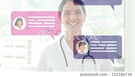 Smiling female physician standing in clinic sporting white coat and stethoscope with chat bubbles Smiling female physician standing in clinic sporting white coat and stethoscope with chat bubbles 135586739
