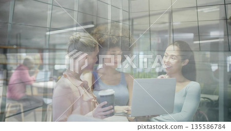 Collaborating three women working on laptop in office lounge, with glass partition and coffee cup 135586784