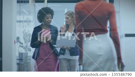 Walking coworkers discussing project in glass-walled office corridor, with laptop and coffee mug 135586790