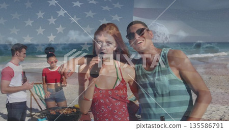 Sipping beer couple in red sundress and striped tank top chatting on sandy beach, with grill 135586791