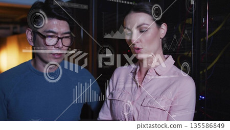 White woman and Asian man studying data visualization on glass panel in server room, with racks 135586849