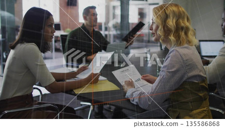Office team sitting around glass conference table, with printed reports folder laptop water glasses 135586868