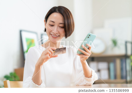 A middle-aged woman looking at her smartphone and credit card in the living room 135586999