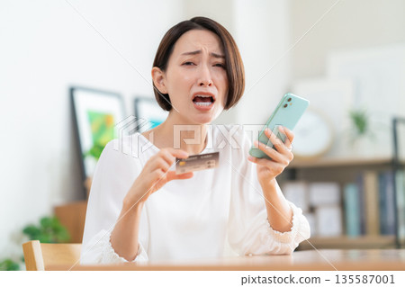 A middle-aged woman looking at her smartphone and credit card in the living room 135587001