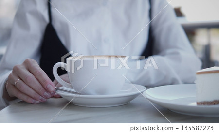 A businesswoman in a white shirt enjoys a cup of coffee at a cafe. She sits casually at a table with a slice of cake beside her, focused and relaxed in the afternoon light. 135587273