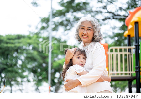 Old woman grandmother patting head kid girl granddaughter as standing hugging in park's playground. 135587947