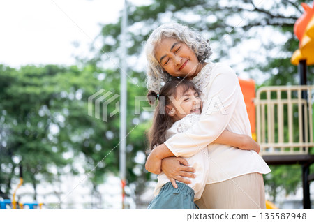 Old woman grandmother and kid girl granddaughter standing hugging each other in park's playground. Old woman grandmother and kid girl granddaughter standing hugging each other in park's playground. 135587948