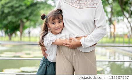 Close up kid granddaughter hugging waist of old woman grandmother standing at railing around river. Close up kid granddaughter hugging waist of old woman grandmother standing at railing around river. 135587967