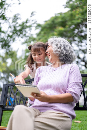 Old woman granny laughing by the drawing in notebook of kid girl granddaughter on bench in the park. Old woman granny laughing by the drawing in notebook of kid girl granddaughter on bench in the park. 135588006