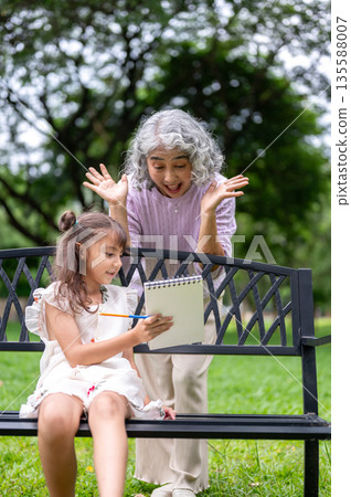 Old woman granny making funny face while looking at notebook that a kid girl granddaughter showing Old woman granny making funny face while looking at notebook that a kid girl granddaughter showing 135588007
