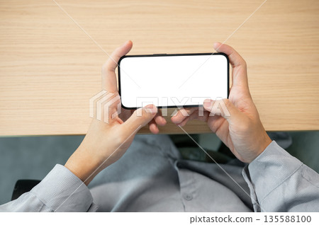 White screen smartphone holding horizontally with both hand while sitting at wooden table in a cafe 135588100