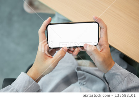 Blank screen smartphone holding horizontally with both hand while sitting at wooden table in a cafe. 135588101