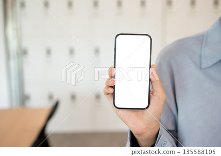 Close up of a man holding white screen smartphone in right hand while standing in cafe or classroom. Close up of a man holding white screen smartphone in right hand while standing in cafe or classroom. 135588105