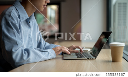 Asian man looking and typing on laptop aside book and coffee while sitting at wooden table in a cafe Asian man looking and typing on laptop aside book and coffee while sitting at wooden table in a cafe 135588167