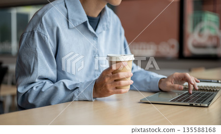 Close up of man holding coffee typing on laptop aside notebook while sitting at wooden table in cafe Close up of man holding coffee typing on laptop aside notebook while sitting at wooden table in cafe 135588168