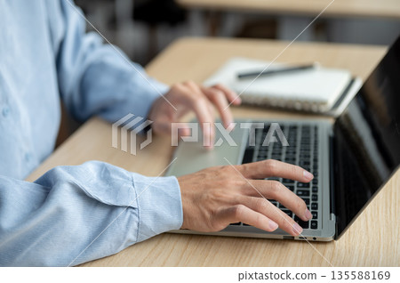 Close up of a man's hands typing on laptop aside notebook while sitting at wooden table in a cafe. Close up of a man's hands typing on laptop aside notebook while sitting at wooden table in a cafe. 135588169