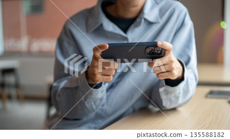 Close up of man looking at phone holding horizontal playing game or watching video at table in cafe. Close up of man looking at phone holding horizontal playing game or watching video at table in cafe. 135588182