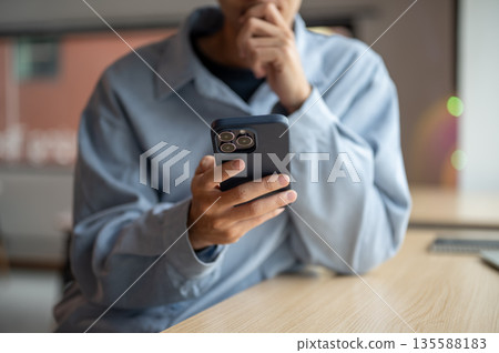 Close up of a man looking at smartphone with hand holding chin while sitting at wooden table in cafe Close up of a man looking at smartphone with hand holding chin while sitting at wooden table in cafe 135588183