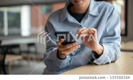 Smiling asian man looking at smartphone with a hand typing while sitting at wooden table in cafe. 135588184