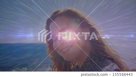 Smiling woman turning toward camera on coastline at sunset, with lens flare streaks over horizon Smiling woman turning toward camera on coastline at sunset, with lens flare streaks over horizon 135588443