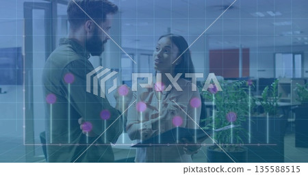 Two colleagues in shirt and blouse discussing data in office, with tablet and stylus, graph overlay 135588515