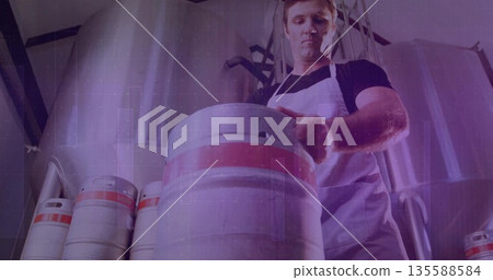 Brewery worker wearing white apron lifting keg on brewery floor, with fermentation tanks, pipes Brewery worker wearing white apron lifting keg on brewery floor, with fermentation tanks, pipes 135588584