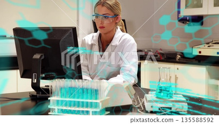 Researcher in white coat and goggles reviewing data on monitor in lab, with test tube rack 135588592