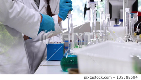 Two technicians in coats gloves using pipette, handling container at bench with flask, copy space Two technicians in coats gloves using pipette, handling container at bench with flask, copy space 135588594