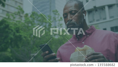 Checking smartphone, man wearing maroon shirt with earbuds in urban park plaza, holding sandwich 135588682