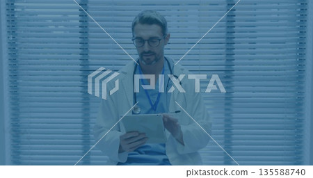 Tapping physician wearing lab coat checking tablet on clinic chair, with stethoscope, eyeglasses 135588740