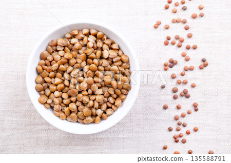 Cooked lentils, boiled small brown lentils, in a white bowl on linen. Canned unshelled mountain lentils, seeds of Lens culinaris, a legume and staple, used for thick curry, gravy or dal. Food photo 135588791
