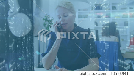 Speaking white executive wearing navy blouse holding smartphone to ear at office, digital overlays 135589411