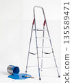 Person uses ladder to paint wall and spills blue paint on floor in indoor space 135589471
