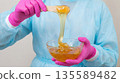 Person in blue protective clothing stirs warm wax in a glass bowl with a wooden stick during a hair removal session 135589482