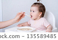 Child learns to eat from a spoon while sitting in a high chair at home during the morning 135589485