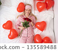 Girl in pink pajamas holds red roses surrounded by heart balloons in a bright bedroom during the morning 135589488