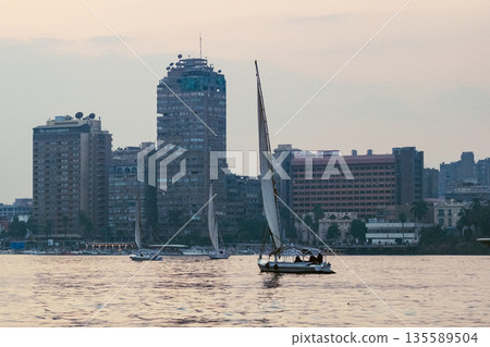 A traditional felucca sailing on the Nile River in Cairo. The boat has a large sail and is surrounded by modern buildings along the riverbank. A traditional felucca sailing on the Nile River in Cairo. The boat has a large sail and is surrounded by modern buildings along the riverbank. 135589504