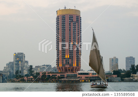 A traditional felucca sailing on the Nile River in Cairo. The boat has a large sail and is surrounded by modern buildings along the riverbank. A traditional felucca sailing on the Nile River in Cairo. The boat has a large sail and is surrounded by modern buildings along the riverbank. 135589508