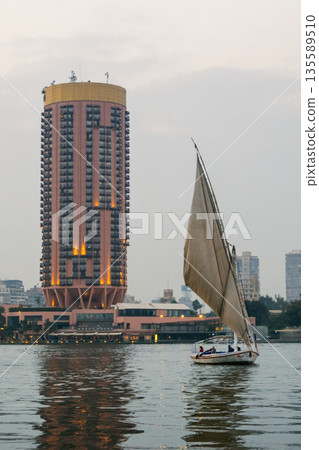 A traditional felucca sailing on the Nile River in Cairo. The boat has a large sail and is surrounded by modern buildings along the riverbank. A traditional felucca sailing on the Nile River in Cairo. The boat has a large sail and is surrounded by modern buildings along the riverbank. 135589510