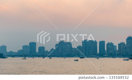 Sunset over the Nile River in Cairo. A tall building stands by the riverbank, with the sun setting behind it, casting warm colors in the sky. Sunset over the Nile River in Cairo. A tall building stands by the riverbank, with the sun setting behind it, casting warm colors in the sky. 135589532