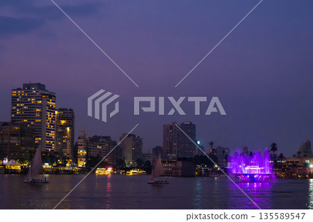 Night view of the Nile River in Cairo. City skyline illuminated by lights. Sailboats on the water. Purple sky with clouds. 135589547