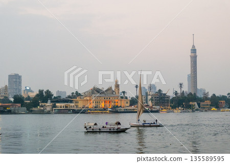 Evening view of the Nile River in Cairo. Two feluccas sail on the water. The background features city buildings and the Cairo Tower. Evening view of the Nile River in Cairo. Two feluccas sail on the water. The background features city buildings and the Cairo Tower. 135589595