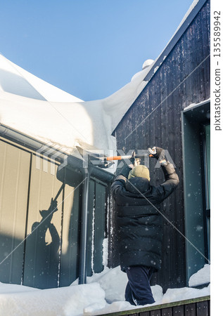 Man clearing heavy snow from a modern house roof on a sunny day. Man clearing heavy snow from a modern house roof on a sunny day. 135589942
