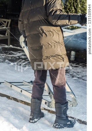 Man in winter clothes carrying a folding ladder in the snow. Man in winter clothes carrying a folding ladder in the snow. 135589944