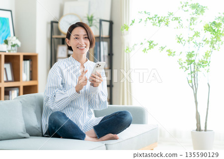 一位中年婦女躺在客廳的沙發上，看著她的智慧型手機。 135590129