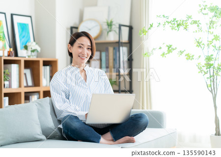 一位中年婦女坐在客廳的沙發上，看著電腦。 135590143