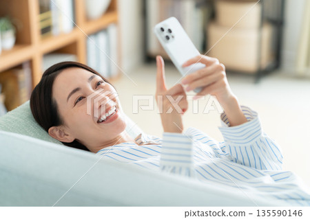 A middle-aged woman lying on the sofa in the living room and looking at her smartphone 135590146